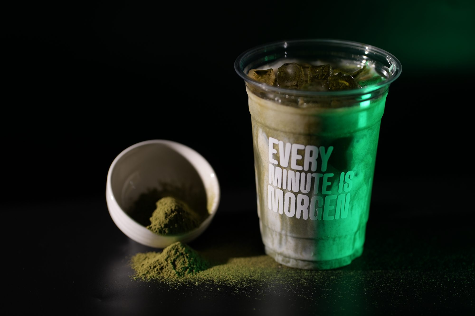 A glowing green iced matcha in an Every Minute is Morgen cup beside a bowl of matcha powder